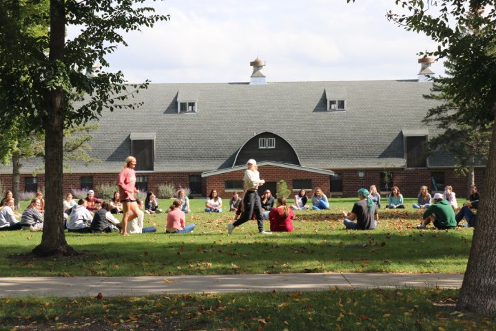 A group of students sitting in a large circle on a grassy lawn, with two people walking in the center. In the background, there is a large barn-style building under a partly cloudy sky, and trees frame the scene.